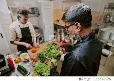 High angle shot of chef sorting fresh lettuce while his sous chef cutting tomatoes in blurred background High angle shot of chef sorting fresh lettuce while his sous chef cutting tomatoes in blurred background 116879829