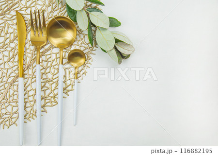 Cutlery: a knife, fork, spoon and a teaspoon of gold color with a white handle lie on a napkin of gold color, next to a green twig on a white background. The concept of food, culture and eating 116882195