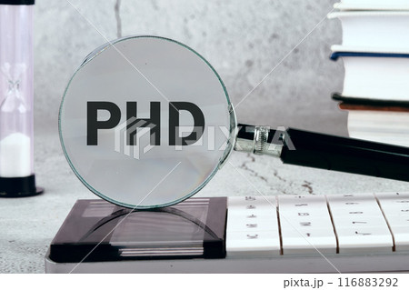 The word PHD is written appeared through a magnifying glass on a gray wall. Photos in black and white 116883292
