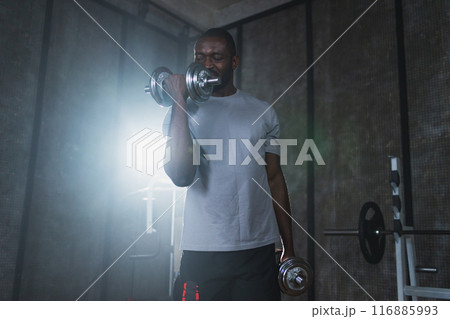 Fitness workout in gym. African American man bodybuilder picking up dumbbells in gym. Workout weight training biceps muscles with dumbbell in fitness club. Man doing sports exercise. Healthy lifestyle 116885993