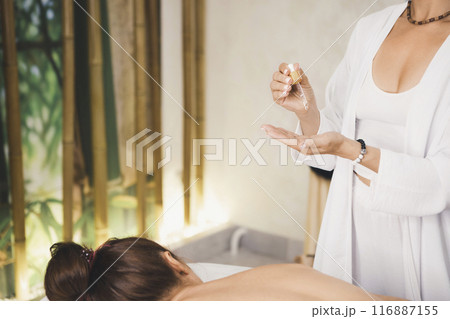 Massage therapist in white outfit takes massage oil before starting work with the client lying on the couch in salon, selective focus. Massage therapist in white outfit takes massage oil before starting work with the client lying on the couch in salon, selective focus. 116887155