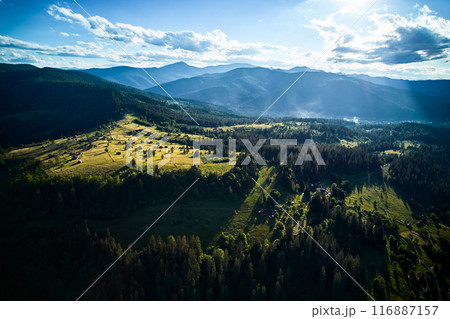 Aerial view of lush, green valley dotted with small houses and winding roads, surrounded by dense forests and rolling hills. Sunlight filters through scattered clouds. Mountains rise under blue sky. 116887157