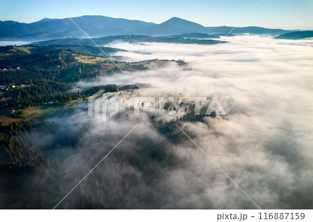 Aerial view reveals misty morning landscape with rolling hills and villages. Golden sunlight breaks through fog, illuminating patches of green and creating dreamy, ethereal scene under clear blue sky. 116887159