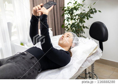 Happy young woman making duck face takes a selfie with a smartphone, lying down on the procedure table, right after a beauty treatment, enjoying the results. Concept of beauty procedures, skin care. 116888280