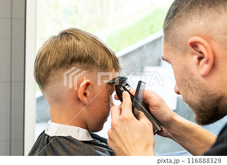 Close-up of man hands grooming kid boy hair in barber shop. Boy cut with hairdresser's machine. Portrait of male child at the barber shop to cut his hair. 116888329