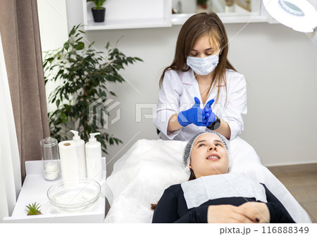 Close-up of beautician using her hands in sterile gloves while applying facial jelly gel to female client's face in beauty clinic. Applying a solution to the face, cosmetic procedure. Close-up of beautician using her hands in sterile gloves while applying facial jelly gel to female client's face in beauty clinic. Applying a solution to the face, cosmetic procedure. 116888349