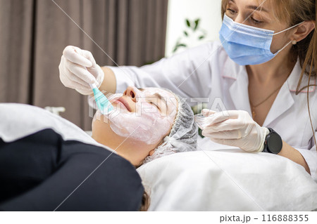 Cosmetologist wearing a surgical mask and gloves, applying a face mask with a spatula to her client 116888355