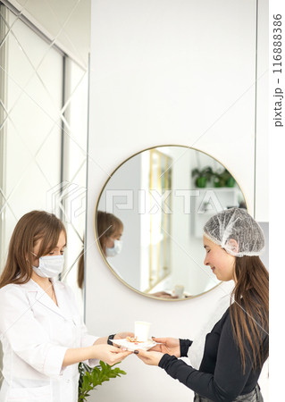 The dermatologist wearing a white suit and a face mask is serving a client with a cup of coffee and some sweets on a serving plate. The client is wearing a disposable cap and a black long sleeve. 116888386