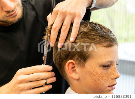 Children hairdresser with scissors is cutting little boy against a dark background. Contented cute preschooler boy getting the haircut. Children hairdresser with scissors is cutting little boy against a dark background. Contented cute preschooler boy getting the haircut. 116888391