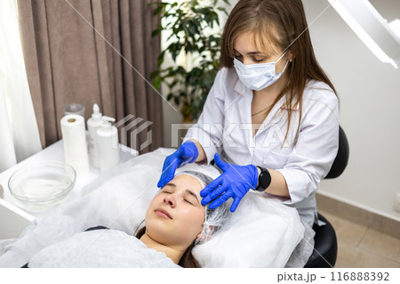 Facial massage is prepared in the beauty salon. Doctor's hands. Treatment for healthy skin. Head of a young woman. Cleansing rejuvenation. Dermatological face mask. Detox therapy Facial massage is prepared in the beauty salon. Doctor's hands. Treatment for healthy skin. Head of a young woman. Cleansing rejuvenation. Dermatological face mask. Detox therapy 116888392