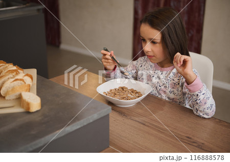 A young girl is seated at a wooden table eating a bowl of cereal for breakfast. Sliced bread is visible on the counter nearby, creating a cozy breakfast setting. A young girl is seated at a wooden table eating a bowl of cereal for breakfast. Sliced bread is visible on the counter nearby, creating a cozy breakfast setting. 116888578