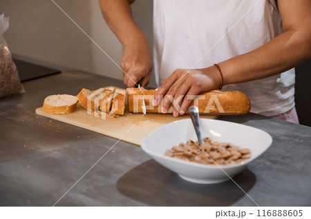 Close-up of a person slicing bread on a cutting board in a modern kitchen, with a bowl of cereal in the foreground. Ideal for breakfast and food preparation concepts. 116888605
