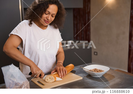 A happy woman in a modern kitchen slicing bread on a cutting board, enjoying the process of cooking. A bowl of cereal is visible on the countertop. 116888610