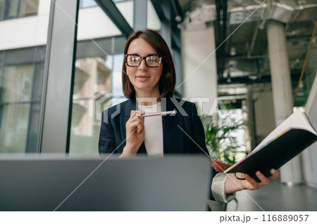 Professional businesswoman with glasses in modern office holding a notebook and pen Professional businesswoman with glasses in modern office holding a notebook and pen 116889057