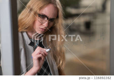 Caucasian woman with glasses writes text in English on a glass wall. Caucasian woman with glasses writes text in English on a glass wall. 116892282