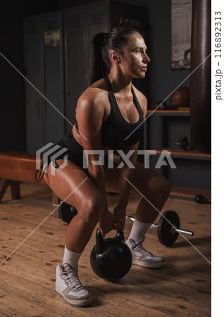 Caucasian woman doing squats with kettlebell in retro gym. Vertical photo.  116892313