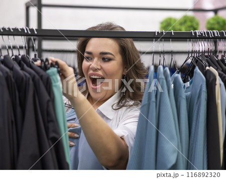 Portrait of a fat woman in a plus size store through hangers with clothes. Portrait of a fat woman in a plus size store through hangers with clothes. 116892320