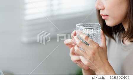 Close-up of a woman holding a glass of water, highlighting hydration and healthy living in a well-lit, modern interior. 116892405