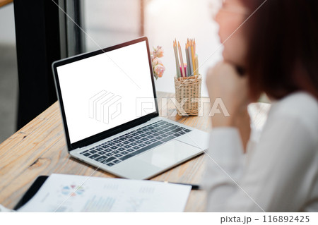 A woman working on a laptop with a blank screen at a wooden desk, surrounded by office supplies and documents, in a modern office environment. 116892425