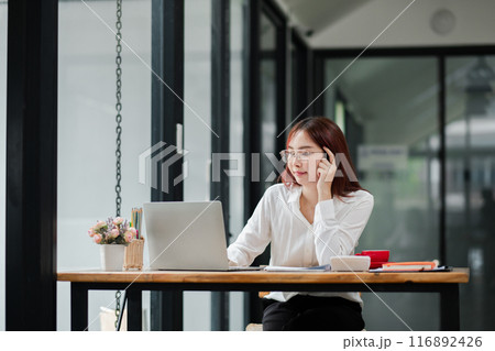 A young woman in a white shirt working on a laptop in a modern office with glass walls, talking on the phone. 116892426