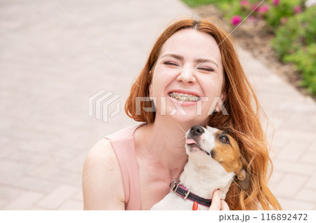 Dog jack russell terrier licks the owner in the face outdoors. Girl with braces on her teeth.  116892472