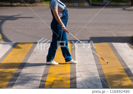 Close-up of the legs of a blind woman crossing the road at a crosswalk with a cane.  116892480