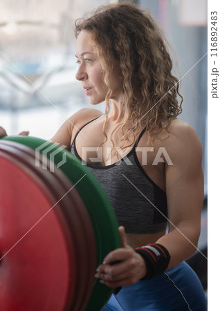 Caucasian forty year old woman putting a weight plate on a barbell in the gym. Vertical photo.  116892483