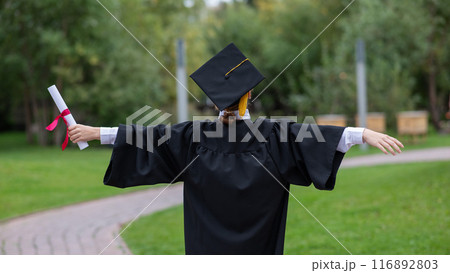 Back view of happy caucasian woman in graduate gown holding diploma outdoors.  116892803
