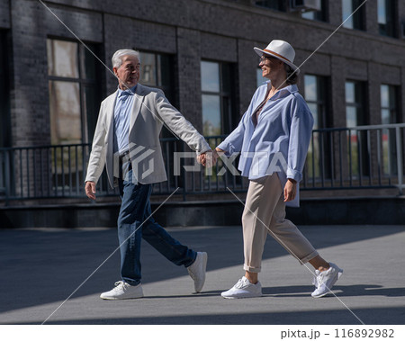 Stylish mature couple. Gray-haired man and woman holding hands while walking around the city. Stylish mature couple. Gray-haired man and woman holding hands while walking around the city. 116892982
