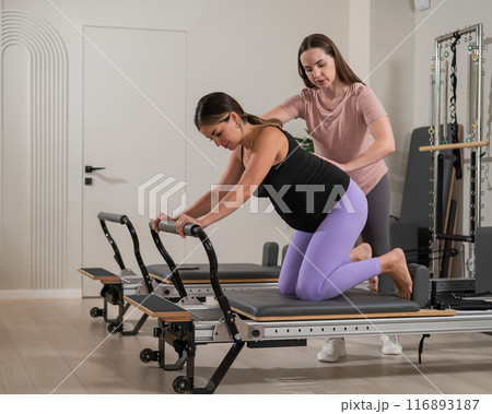 A pregnant woman works out on a reformer exercise machine with a personal trainer. A pregnant woman works out on a reformer exercise machine with a personal trainer. 116893187