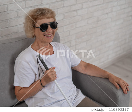 An elderly blind woman wearing sunglasses and with a tactile cane sits on the sofa. An elderly blind woman wearing sunglasses and with a tactile cane sits on the sofa. 116893357