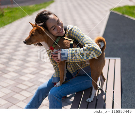 Young beautiful woman hugging a dog while sitting on a bench outdoors. Non-barking African basenji dog. Young beautiful woman hugging a dog while sitting on a bench outdoors. Non-barking African basenji dog. 116893449