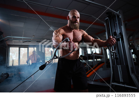 Bald Caucasian bodybuilder training chest using cable crossover in gym.  116893555
