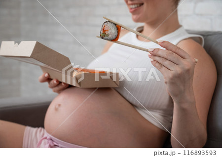 A pregnant woman sits on the sofa and eats rolls from a box. Food delivery.  116893593