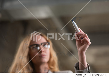 Caucasian woman with glasses writes text in English on a glass wall.  116893657