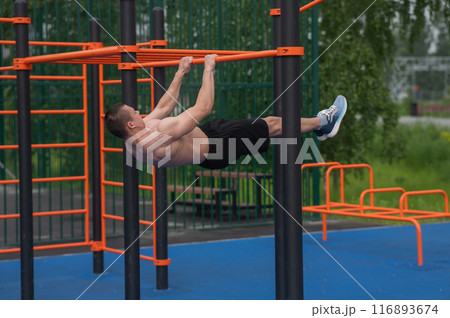 Shirtless male athlete doing front hang on horizontal bar outdoors.  116893674