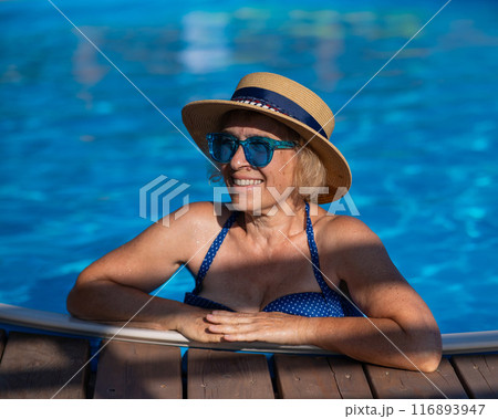Portrait of an elderly woman in a hat and blue sunglasses swims in the pool. Vacation in retirement.  116893947