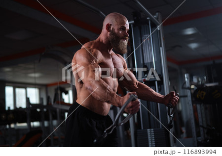Bald Caucasian bodybuilder training chest using cable crossover in gym.  116893994