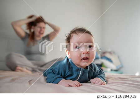 A three-month-old boy lies on his stomach on the bed and his mother sits behind him and cries. Postpartum depression. A three-month-old boy lies on his stomach on the bed and his mother sits behind him and cries. Postpartum depression. 116894019