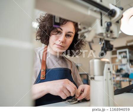 A woman tanner sews a leather belt on a sewing machine.  116894059