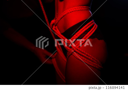 Man tightens ropes using Japanese shibari technique on woman's body in red light.  116894141