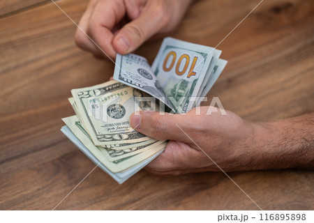 Male company accountant holds dollar banknotes of different denominations carefully counting cash for weekly income report at end of week sitting at wooden table in office 116895898