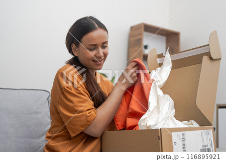 Fashionable Joy: On her couch, a cheerful lady unboxes her parcel, thrilled with her successful online fashion store shopping and express delivery.  116895921