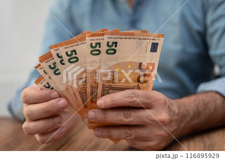 Male financial manager holds euro banknotes fan in hands carefully counting money for income report in end of working day sitting at wooden desk in company office Male financial manager holds euro banknotes fan in hands carefully counting money for income report in end of working day sitting at wooden desk in company office 116895929