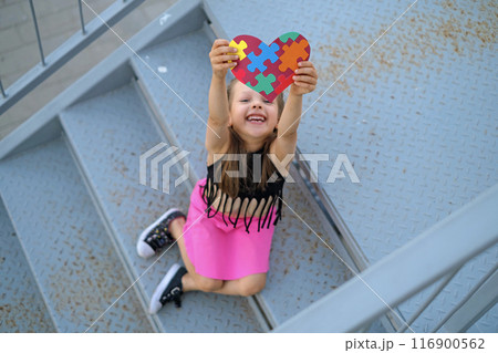 Funny laughing adorable girl shows a heart card made of colorful puzzles as a sign of support for children with autism spectrum disorder 116900562