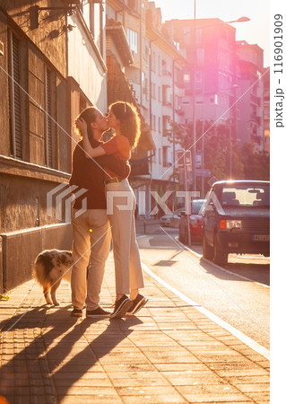 Romantic couple kissing on the street. Family walking with dog pet. Sunset light in background. Vertical orientation Romantic couple kissing on the street. Family walking with dog pet. Sunset light in background. Vertical orientation 116901909
