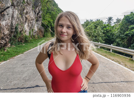 Young woman posing confidently on road surrounded by lush green nature under cloudy sky Young woman posing confidently on road surrounded by lush green nature under cloudy sky 116903936