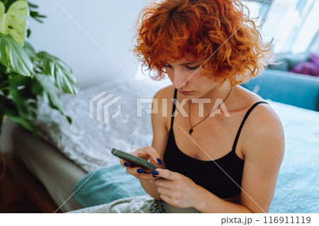 portrait of young woman with curly red hair sitting on bed in bedroom using phone 116911119