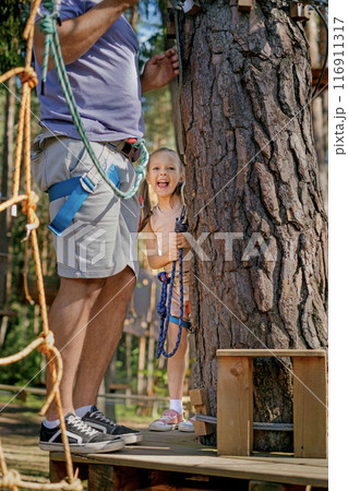 A funny child in full protective gear walks a rope route at a height with his dad A funny child in full protective gear walks a rope route at a height with his dad 116911317