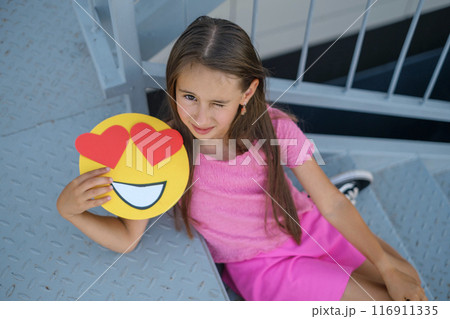 A winking Girl sitting on the stairs shows emojis with loving eyes and big hearts A winking Girl sitting on the stairs shows emojis with loving eyes and big hearts 116911335